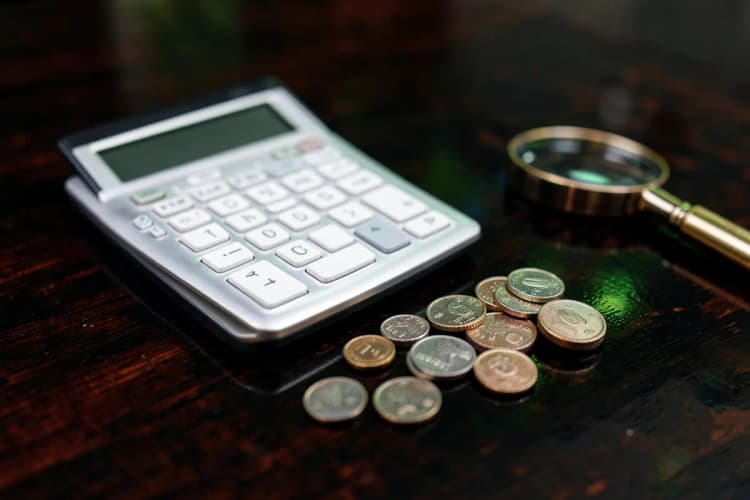 A close-up of a sleek, modern calculator placed next to a small stack of coins and a magnifying glass on a dark wooden surface.